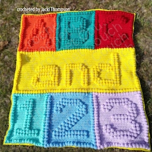 Peut inclure: Une couverture pour bébé en crochet avec le mot "ONE" en fil jaune et les chiffres "1" et "2" en fil bleu et violet. La couverture est réalisée avec un point texturé qui crée un effet 3D.