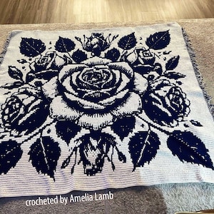 Peut inclure: Une couverture crochetée bleu marine et blanche avec un grand motif de roses. La couverture a une bordure à franges et le texte "crocheted by Amelia Lamb" est visible. Le motif floral comprend des roses et des feuilles.