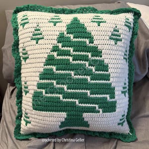 Op de afbeelding: Een wit gehaakt kussen met een groen kerstboomontwerp. Het kussen heeft een witte gehaakte rand met een gegolfde rand. De tekst "crocheted by Christina Geller" staat onderaan het kussen.