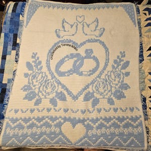 Könnte beinhalten: Eine rechteckige Häkeldecke in Weiß und Hellblau mit einem zentralen Herzmotiv, das zwei ineinander verschlungene Ringe enthält. Über dem Herz befinden sich zwei Tauben und ein kleines Herz. Darunter sind Rosen und der Text "crocheted by Terena White".