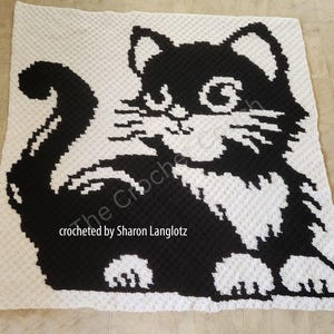May include: Black and white crochet blanket featuring a stylized cat design. The cat is sitting with its tail curled around its body. The blanket is crocheted in a textured stitch.