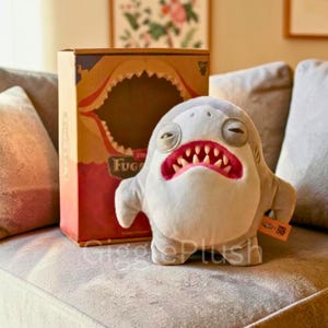 Op de afbeelding: Een lichtgrijze pluche haai met een brede, tandige grijns en slaperige ogen. De haai staat naast een kartonnen doos met een haaienbekontwerp en het woord "Fug". Het speelgoed heeft een klein label.