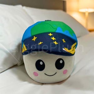 May include: A soft toy with a smiling face, black eyes, and pink cheeks. It is wearing a blue and green cap designed to look like the Earth, with yellow stars and accents. The toy is on a white surface.
