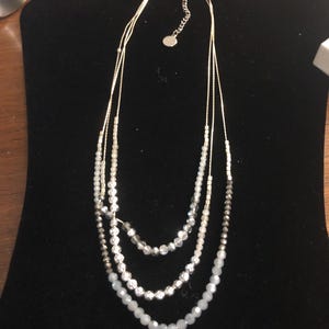Puede incluir: Collar de tres hilos con cuentas blancas, plateadas y grises. El collar se exhibe en un expositor de joyer&iacute;a de terciopelo negro. Las cuentas var&iacute;an en tama&ntilde;o y textura, creando un efecto de capas.