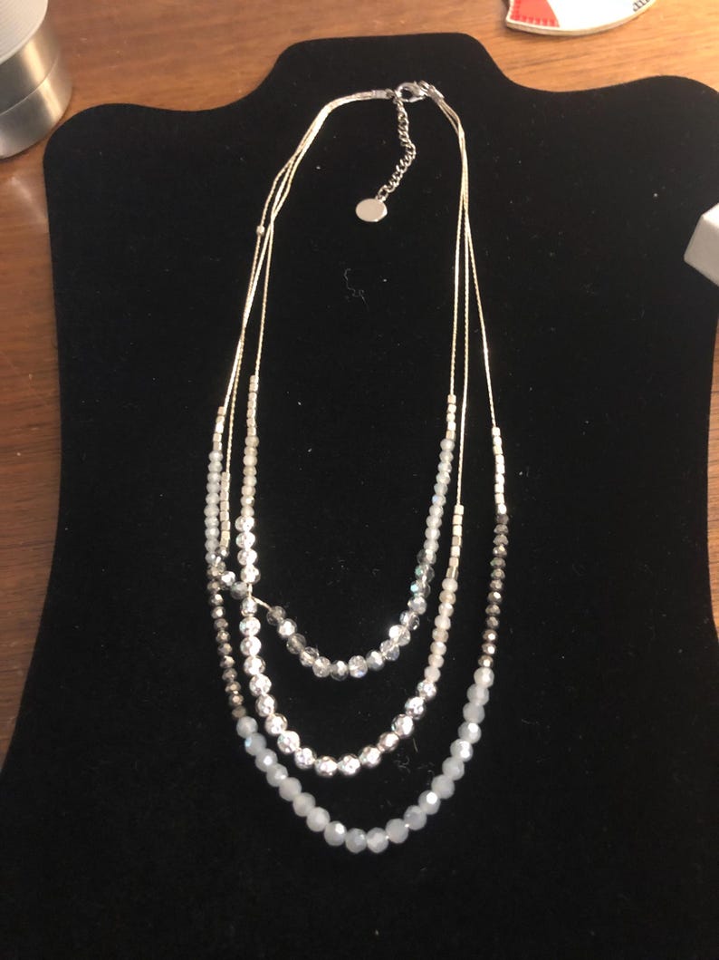 Puede incluir: Collar de tres hilos con cuentas blancas, plateadas y grises. El collar se exhibe en un expositor de joyer&iacute;a de terciopelo negro. Las cuentas var&iacute;an en tama&ntilde;o y textura, creando un efecto de capas.