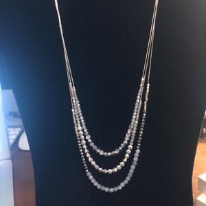 Puede incluir: Un collar de m&uacute;ltiples hilos con cadenas y cuentas plateadas. El collar presenta tres capas de diferentes tama&ntilde;os y colores de cuentas, que van desde el transparente hasta el gris y la plata. El collar se exhibe en un maniqu&iacute; negro.