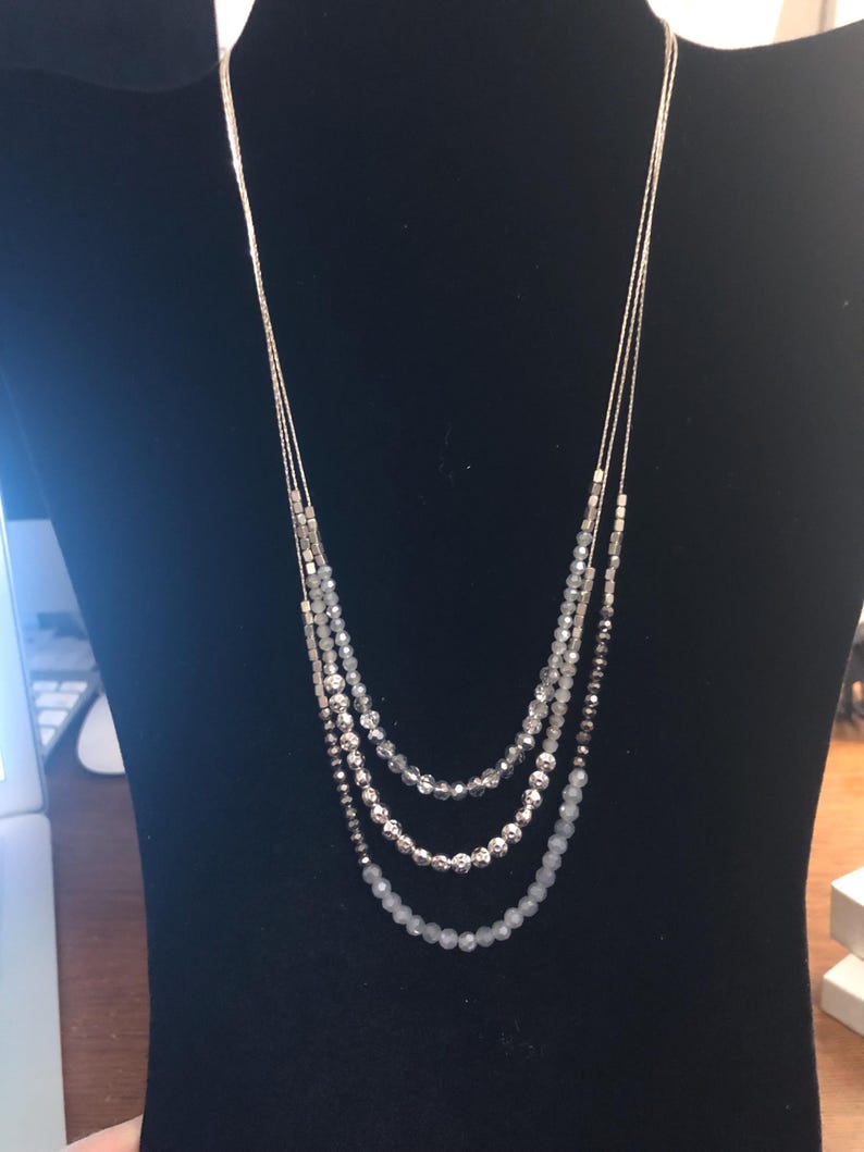 Puede incluir: Un collar de m&uacute;ltiples hilos con cadenas y cuentas plateadas. El collar presenta tres capas de diferentes tama&ntilde;os y colores de cuentas, que van desde el transparente hasta el gris y la plata. El collar se exhibe en un maniqu&iacute; negro.