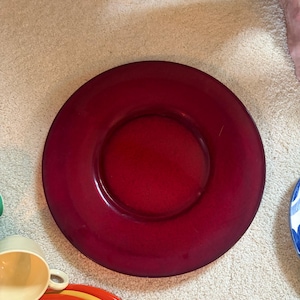 May include: A large, circular, deep red plate with a smooth, glossy finish and a wide, flat rim. The plate features a central recessed area and is positioned alongside a stack of colorful plates and a light yellow cup in the lower corner.