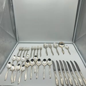 May include: A collection of silver-colored flatware, including forks, spoons, and knives, arranged on a white surface. The spoons vary in size, and the knives have a decorative handle design. The set appears to be for a formal dining setting.