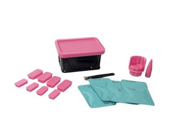 Può includere: Un contenitore nero con coperchio rosa, otto piccoli blocchi rettangolari rosa, quattro bustine azzurre con testo scritto a mano, un utensile nero, una piccola sedia rosa e un oggetto appuntito rosa su sfondo bianco.