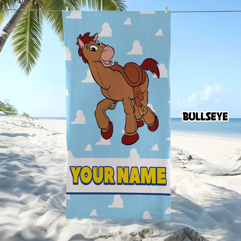 Op de afbeelding: Een rechthoekige strandhanddoek met een lichtblauwe achtergrond en witte wolkenpatronen. Een bruin cartoonpaard met een zadel staat in het midden, met de tekst BULLSEYE erboven en de tekst YOUR NAME in het geel onderaan.