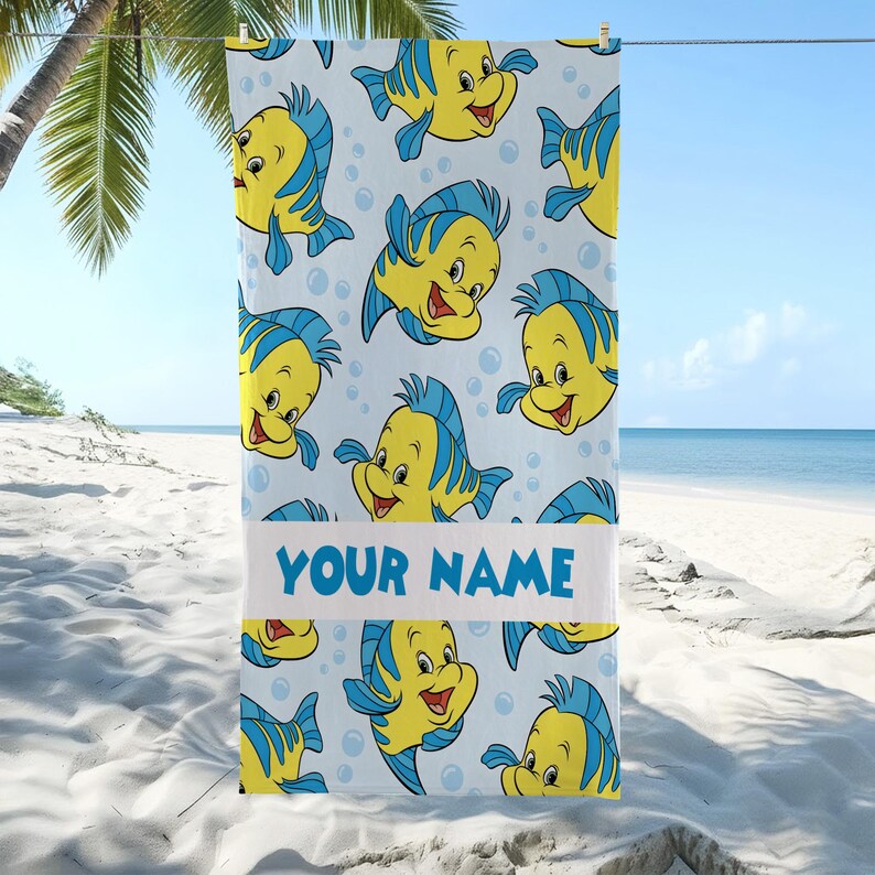 May include: A rectangular beach towel featuring a repeating pattern of yellow and blue cartoon fish on a light blue background with bubbles. A horizontal white band across the center displays the blue text YOUR NAME in a bold sans-serif font.