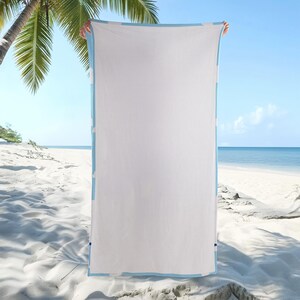 Puede incluir: Una toalla de playa rectangular que presenta un panel central grande de color blanco s&oacute;lido enmarcado por un borde azul claro. La toalla se sostiene verticalmente, mostrando su dise&ntilde;o limpio y minimalista y su textura suave en un entorno luminoso.