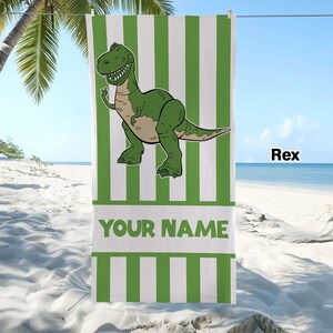 May include: A rectangular beach towel featuring vertical green and white stripes. A green cartoon dinosaur is centred in the upper portion. Below the dinosaur, a white horizontal band contains the green text YOUR NAME in capital letters.