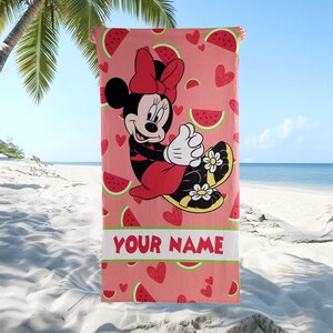 Pu&ograve; includere: Un telo mare rettangolare con sfondo rosa e un motivo ripetuto di fette di anguria rosse e piccoli cuori rossi. Minnie Mouse &egrave; al centro, indossa un fiocco rosso e sandali gialli. Il testo YOUR NAME &egrave; stampato in colore rosso.