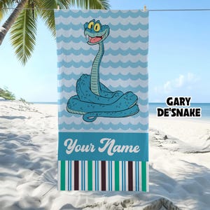 May include: A rectangular beach towel featuring a cartoon blue snake with large yellow eyes, set against a light blue wavy pattern. The text GARY DE'SNAKE appears on the right, with the words Your Name and vertical stripes at the bottom.