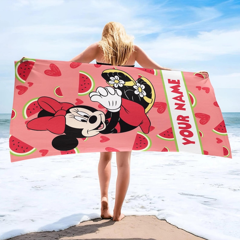 Pu&ograve; includere: Un telo mare rettangolare con sfondo rosa decorato da cuori rossi e fette di anguria. Minnie Mouse &egrave; raffigurata al centro con un fiocco rosso e sandali gialli. Una striscia verticale bianca riporta la scritta YOUR NAME.