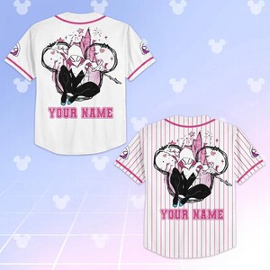 May include: White baseball jersey with pink accents and a cartoon graphic of a superhero. The jersey features the text "YOUR NAME" in pink. The jersey has short sleeves and a button-up front.
