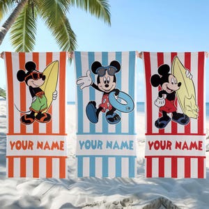 May include: Three rectangular beach towels featuring Mickey Mouse graphics. The left towel is orange-striped, the center is blue-striped, and the right is red-striped. Each towel displays the text YOUR NAME at the bottom in a bold font.