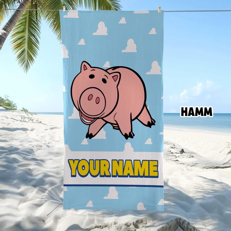 Puede incluir: Una toalla de playa azul claro con un patr&oacute;n de nubes blancas, una ilustraci&oacute;n central de un cerdo rosa, el texto HAMM en negro y el texto YOUR NAME en letras amarillas en negrita con un borde azul oscuro en la parte inferior.