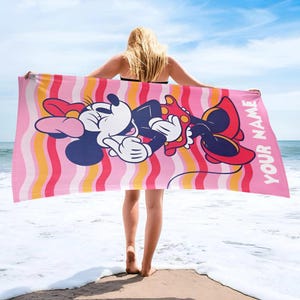 May include: A rectangular beach towel featuring a graphic of Minnie Mouse wearing a red polka dot dress and bow. The background has vertical wavy stripes in shades of pink, orange, and white. The text YOUR NAME is printed in white on the right side.