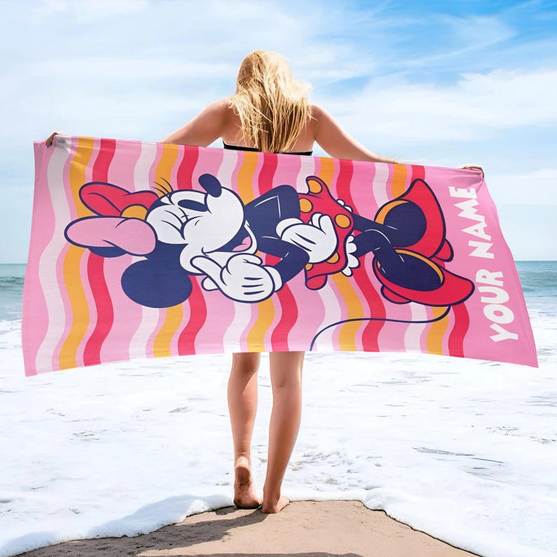May include: A rectangular beach towel featuring a graphic of Minnie Mouse wearing a red polka dot dress and bow. The background has vertical wavy stripes in shades of pink, orange, and white. The text YOUR NAME is printed in white on the right side.