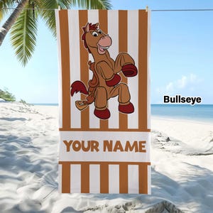 May include: A rectangular beach towel featuring vertical brown and white stripes. In the centre, a cartoon brown horse with a saddle and dark red hooves is depicted. Below the horse, a white horizontal band contains the text YOUR NAME.