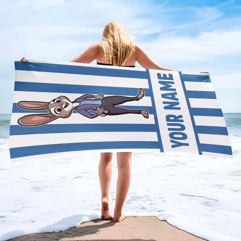 Puede incluir: Una toalla de playa rectangular con un patr&oacute;n de rayas verticales azules y blancas. La toalla muestra un personaje de conejo de dibujos animados con una chaqueta morada y pantalones oscuros, con el texto YOUR NAME en letras azules.