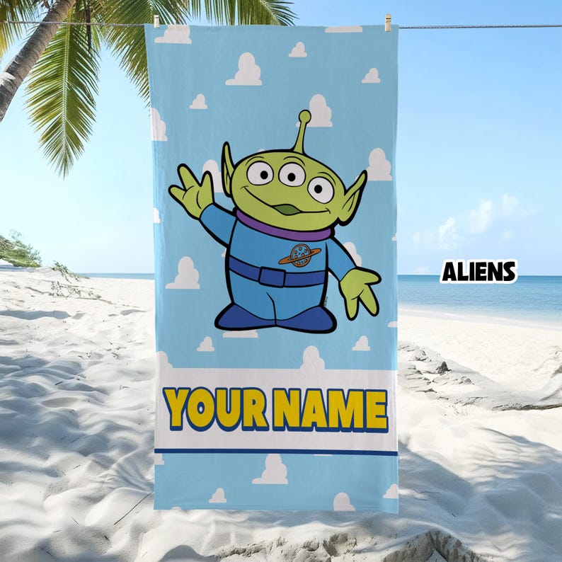 Op de afbeelding: Een lichtblauwe strandhanddoek met een herhalend patroon van witte wolken en een centrale afbeelding van een groen buitenaards wezen met drie ogen in een blauw ruimtepak. De tekst "YOUR NAME" staat in grote gele letters onderaan de handdoek.