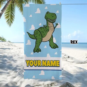 Puede incluir: Una toalla de playa rectangular con fondo azul claro y patrones de nubes blancas. Un dinosaurio de dibujos animados verde est&aacute; centrado en la toalla. Debajo del dinosaurio, el texto YOUR NAME est&aacute; impreso en letras amarillas con borde azul.