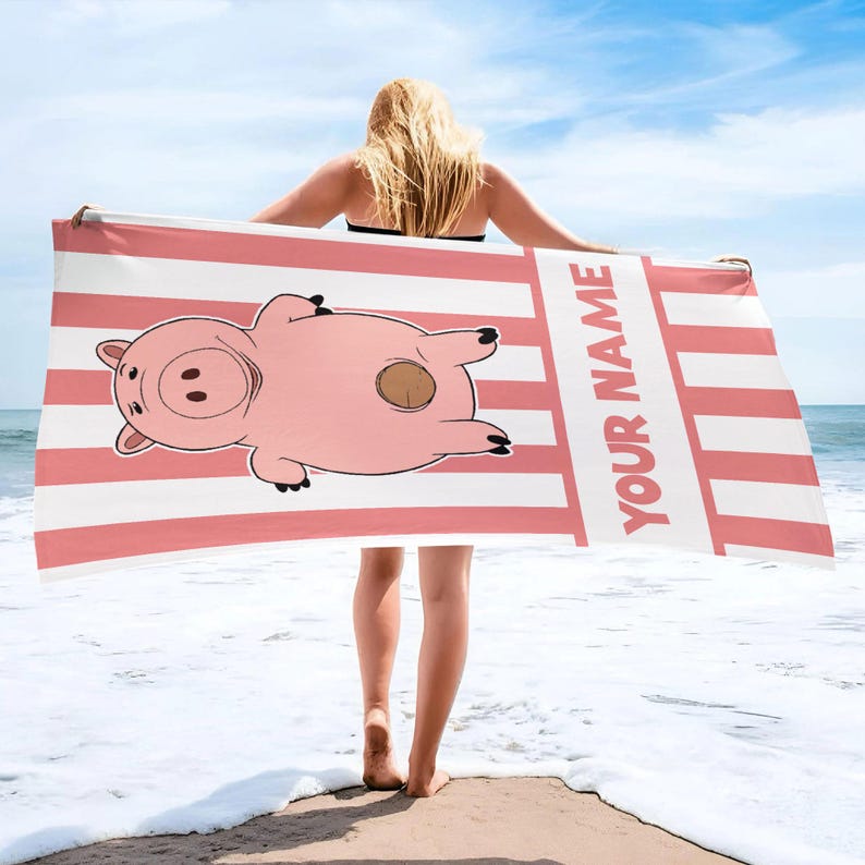 May include: A rectangular beach towel featuring a pink and white vertical striped pattern. The towel displays a cartoon illustration of a pink pig lying on its back and the text "YOUR NAME" printed in large, bold, pink capital letters.