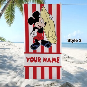 May include: A rectangular beach towel featuring vertical red and white stripes, a graphic of Mickey Mouse holding a yellow surfboard, and the text YOUR NAME in red letters. The towel is displayed hanging on a line with the text Style 3 visible.