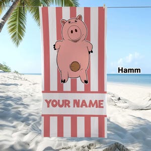 May include: A rectangular beach towel featuring a pink cartoon pig character with a brown circular coin slot on its belly. The towel has vertical pink and white stripes and includes the text "YOUR NAME" in bold red letters and "Hamm" in black.