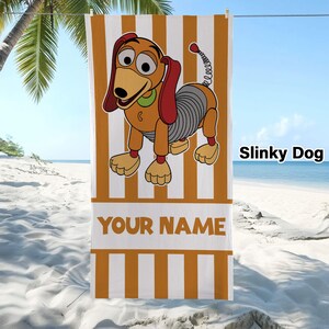 May include: A rectangular beach towel featuring a vertical orange and white striped pattern. The centre displays a cartoon Slinky Dog character with a brown body, red ears, and a silver spring midsection. The text YOUR NAME appears in orange.