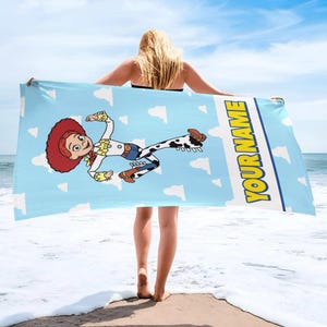 Puede incluir: Una toalla de playa azul claro con un patr&oacute;n de nubes blancas, una ilustraci&oacute;n gr&aacute;fica del personaje Jessie con cabello rojo y traje de vaquera, y el texto amarillo YOUR NAME impreso verticalmente en el lado derecho.