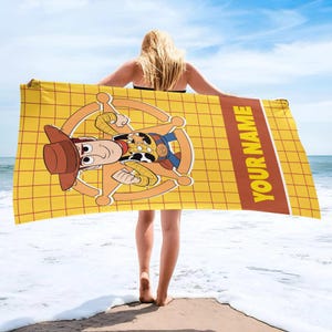 May include: A rectangular beach towel featuring a yellow grid pattern background with a central graphic of a cartoon cowboy character wearing a brown hat and a sheriff badge. A brown vertical stripe on the right side contains the text YOUR NAME.
