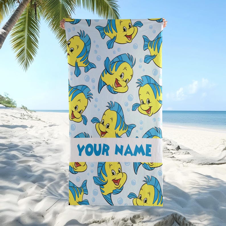 May include: A rectangular beach towel featuring a repeating pattern of yellow and blue cartoon fish on a white background with light blue bubbles. A horizontal white band across the center displays the blue text YOUR NAME in capital letters.