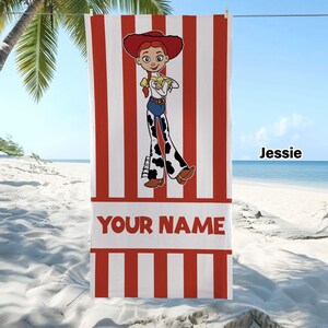 May include: A rectangular beach towel featuring a red and white vertical striped pattern. The centre displays a cartoon character with red hair, a red hat, and cow-print trousers. The text YOUR NAME is printed in red block letters near the bottom.
