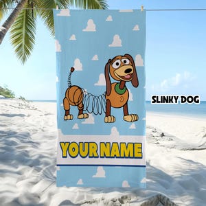 Op de afbeelding: Een lichtblauwe strandhanddoek met een herhalend wit wolkenpatroon en een afbeelding van Slinky Dog. Het personage heeft bruine oren, een groene halsband en een lichaam van een metalen veer. De tekst SLINKY DOG en YOUR NAME staat erop.