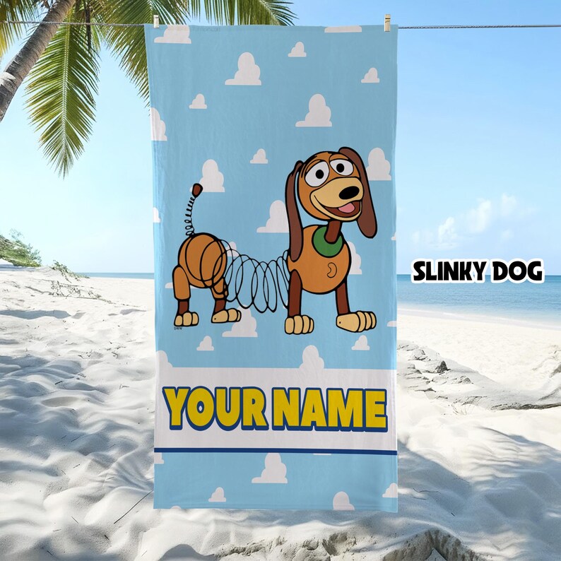 Op de afbeelding: Een lichtblauwe strandhanddoek met een herhalend wit wolkenpatroon en een afbeelding van Slinky Dog. Het personage heeft bruine oren, een groene halsband en een lichaam van een metalen veer. De tekst SLINKY DOG en YOUR NAME staat erop.