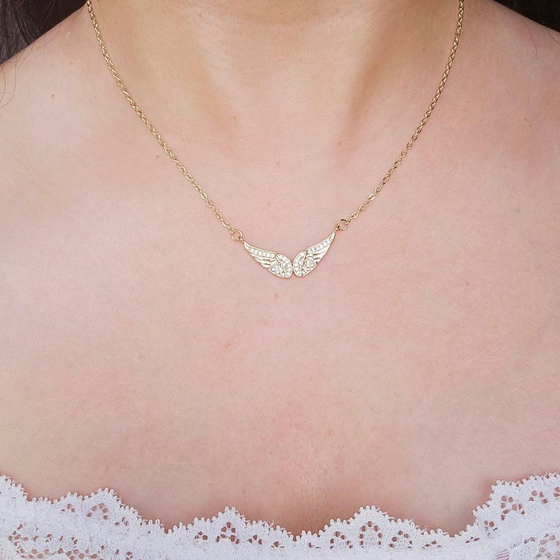 May include: A gold-colored necklace featuring a pair of angel wings, each wing adorned with small, sparkling crystals. The necklace is delicate and hangs on a thin chain, resting on a person's chest.