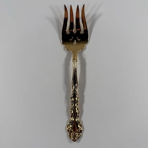 May include: A gold-colored serving fork with four tines and an ornate handle. The fork is designed with a decorative pattern and is likely used for serving food. The overall design suggests a vintage or antique style.