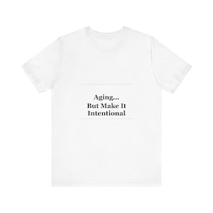 May include: White t-shirt featuring the text "Aging... But Make It Intentional" in a dark, sans-serif font. The text is centered, with a horizontal line above and below the text.