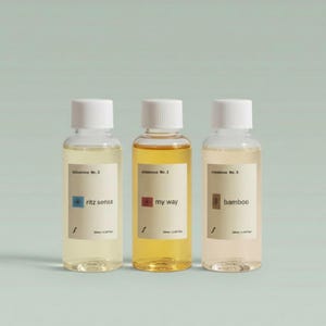 May include: Three small, clear glass bottles with white caps, each containing a different colored liquid. The bottles have labels that read "ritz sense", "my way", and "bamboo". The background is a light teal color.