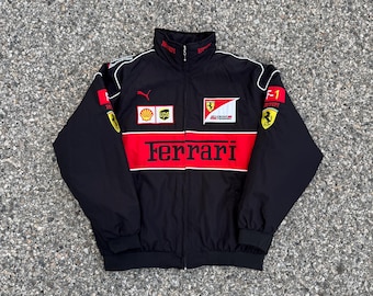 Könnte beinhalten: Eine schwarz-rote Reißverschlussjacke mit Ferrari-Logo, Scuderia Ferrari-Schriftzug und verschiedenen Sponsoren-Aufnähern wie Shell, UPS und Bridgestone. Die Jacke hat einen hohen Kragen, lange Ärmel mit elastischen Bündchen und einen Ferrari-Schriftzug.