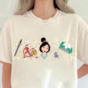 May include: Cream-colored t-shirt featuring a graphic design of characters and symbols from a popular animated film. The design includes a sword, a dragon, a woman, a cricket, and a hair comb, all in vibrant colors. The shirt has short sleeves and a crew neck.