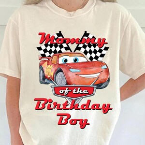 May include: Cream-colored t-shirt featuring a red cartoon race car with the number 95. The shirt has the text "Mommy of the Birthday Boy" in red. Two checkered flags are above the car.