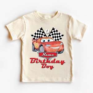May include: A cream-colored t-shirt featuring a red cartoon race car with the number 95. Above the car are two checkered flags. The shirt has the words "Name Birthday Boy" in red script.