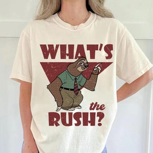 May include: Cream-colored t-shirt featuring a cartoon sloth in a green shirt, brown pants, and a red and blue striped tie. The text "WHAT'S the RUSH?" is printed in a distressed red font.