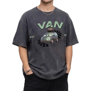 May include: Dark gray t-shirt featuring a cartoon van with the word "VAN" in light green above it. The van has a luggage carrier on top and is set against a checkered flag design. The text "EST 19" is also visible.